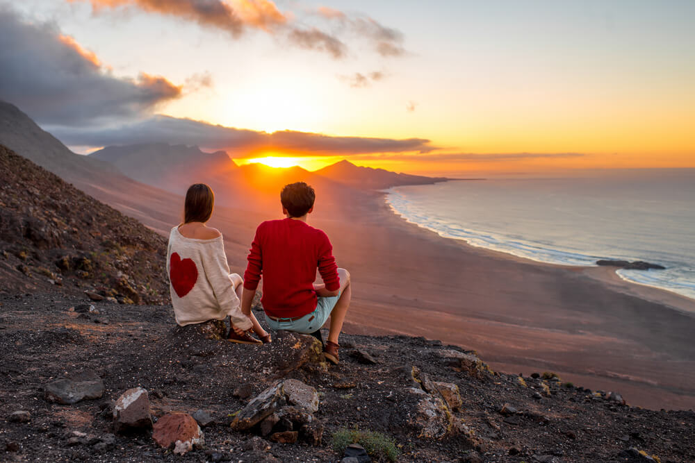 Giovane coppia che gode di un bel tramonto seduti insieme sulla montagna con una splendida vista sulla costa di Cofete sull'isola di Fuerteventura